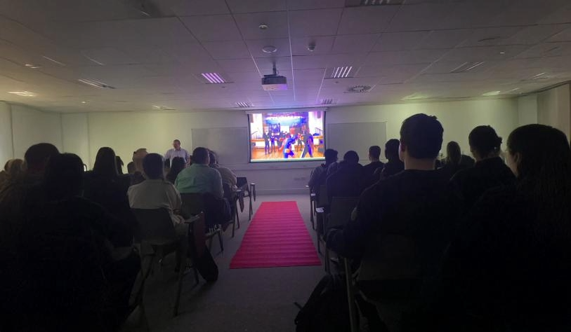Group of TY students sitting in dimmed lite room with red carpet rolled out on floor, watching film on screen during awards ceremony