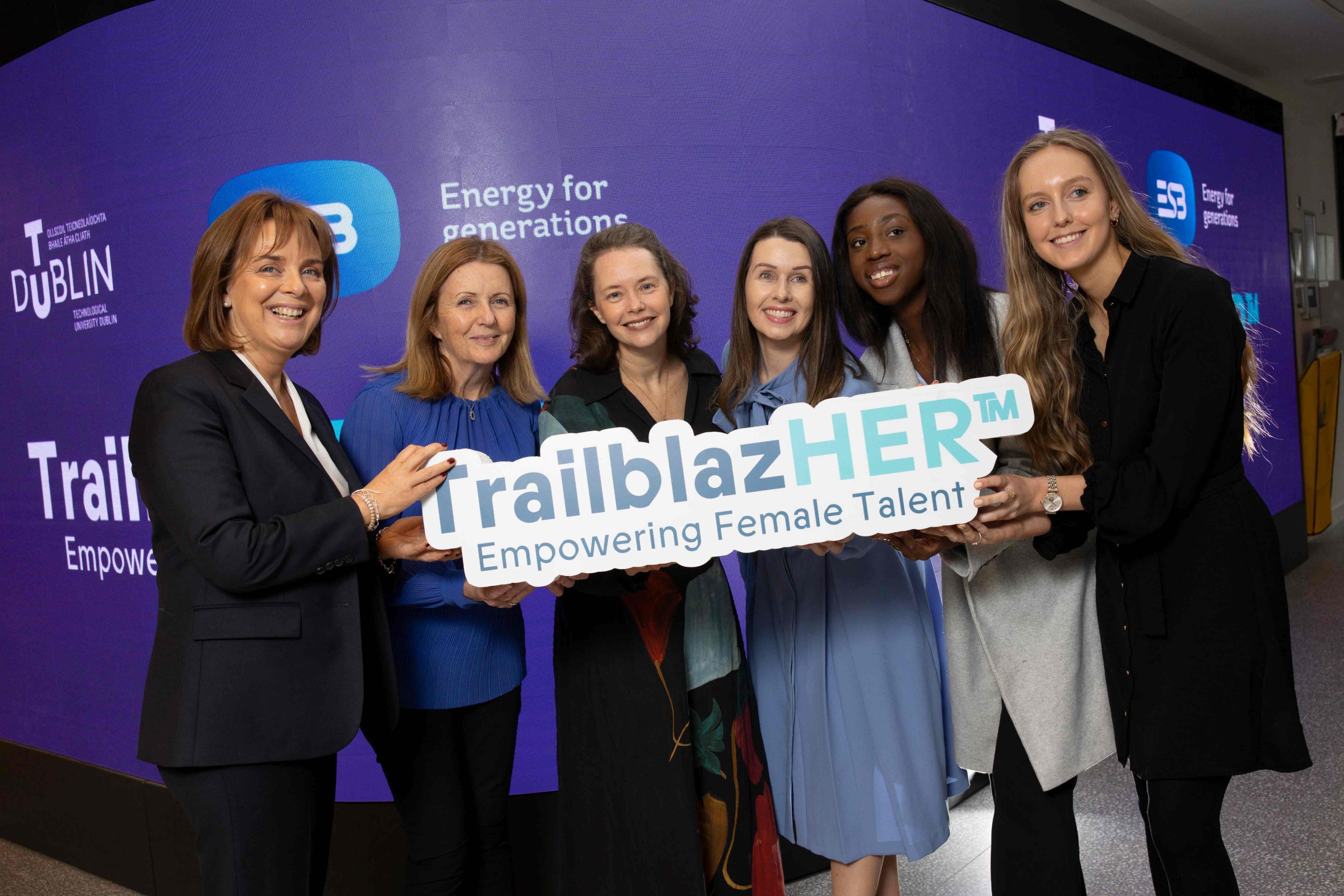 A group of women hold a TailblazHER sign in front of a large purple screen