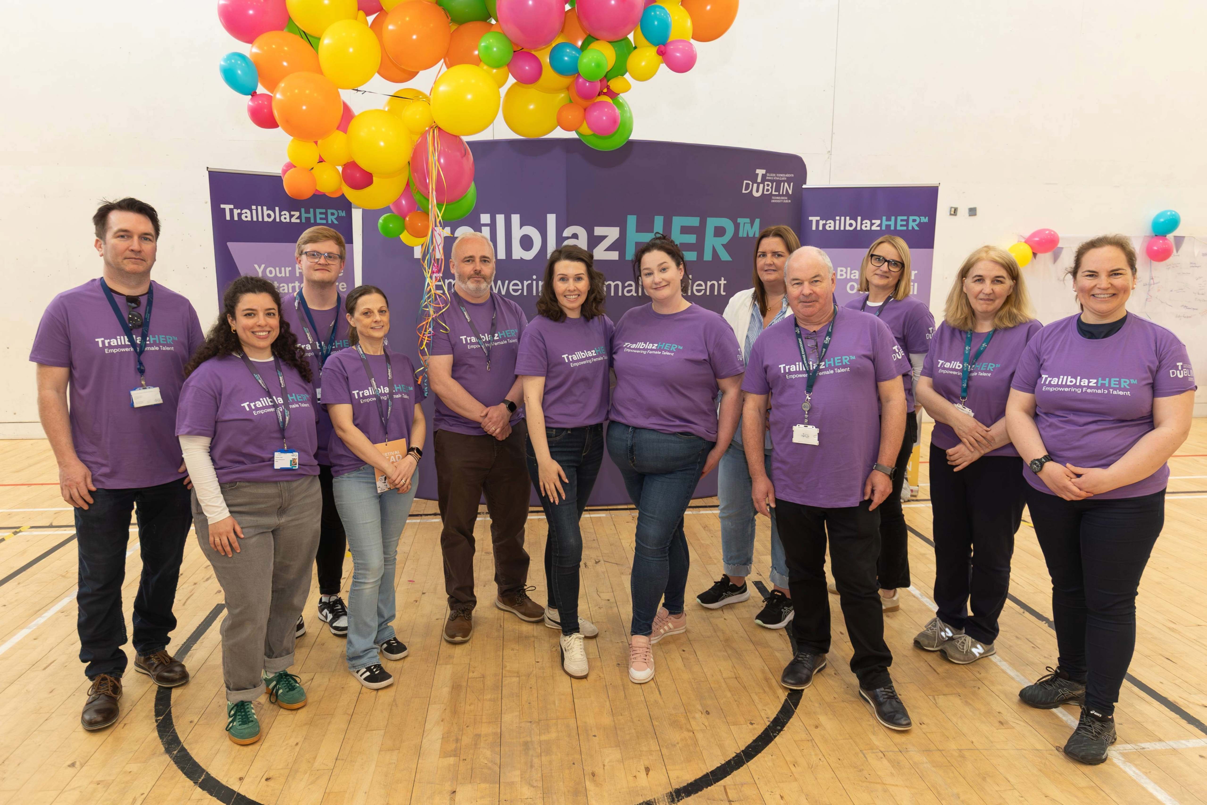 A group of people in purple TrailblazHER™ t-shirts pose in front of a branded stand with pullups on either side and a bunch of colourful balloons in the top left.