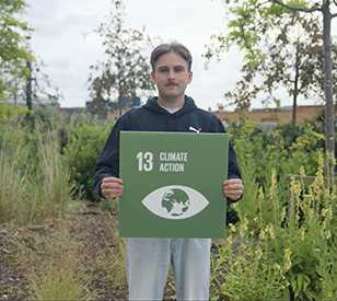 Young white man holding a square green sign with SDG13 Climate Action against backdrop of plants and foliage