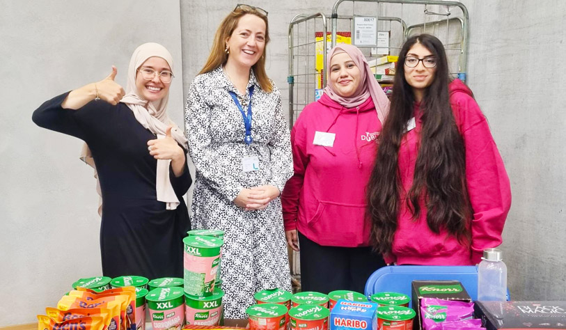three students, two in fuchsia pink hoodies, and a staff giving out free food at a food pantry table