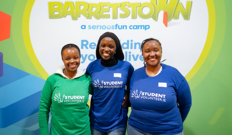 3 female student volunteers smile for camera in front of Barrettstown Charity Poster Backdrop