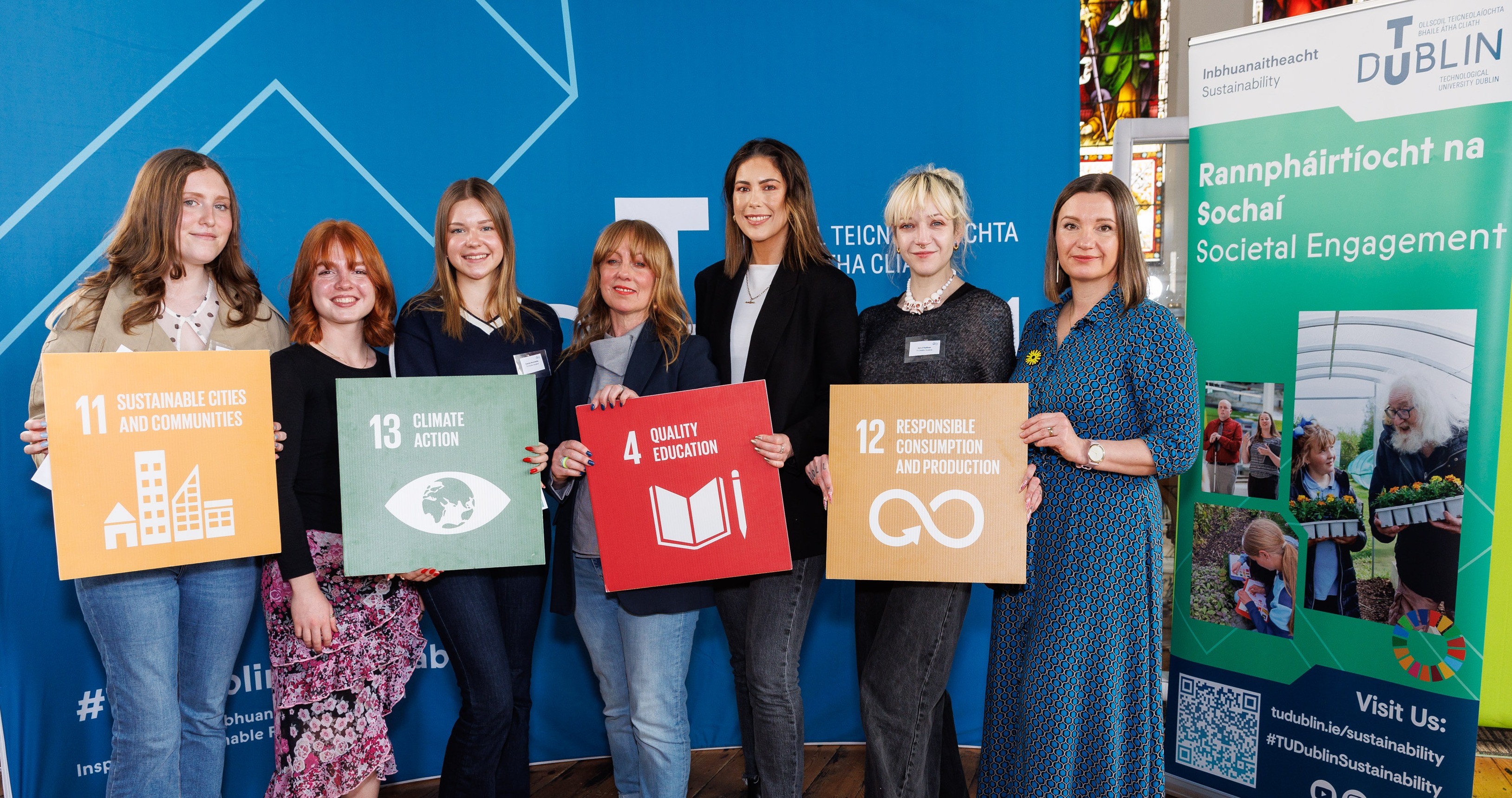 Five women holding signs depicting sustainable development goals, in front of TU Dublin signag