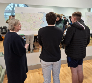 one woman and three young men stand in front of large map. they face away from us. there are others in the background which can be seen as one wall is mirrored.