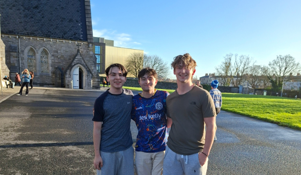 three male students smile for camera in campus backdrop of grey church building, blue sky and green grass