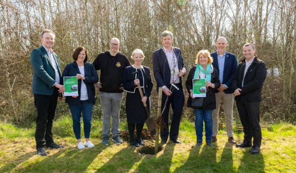 three white women and five white men smile for camera. woman in centre holds new tree and man in centre holds shovel