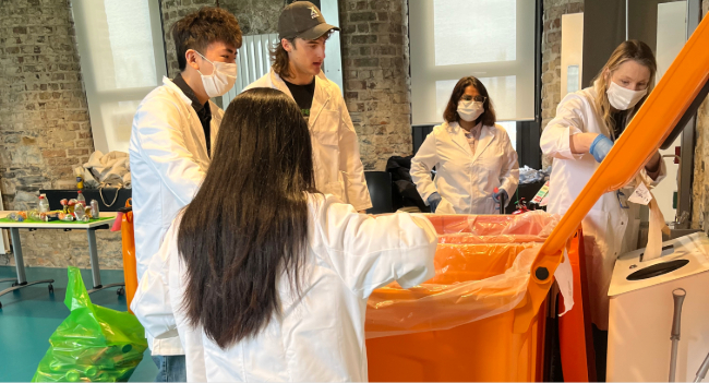 2 males 3 females in white coats taking litter out of large orange bin