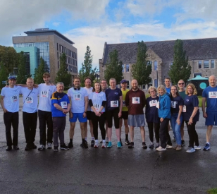 group of 20 people wearing running gear and race numbers pose for camera against campus backdrop with mixture of old and new buildings
