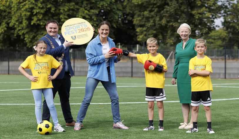 A group of adults and children pose on a sports field for Dublin City Council SportsFest 2026, with one adult holding a SportsFest sign and another holding boxing gloves with a young participant.