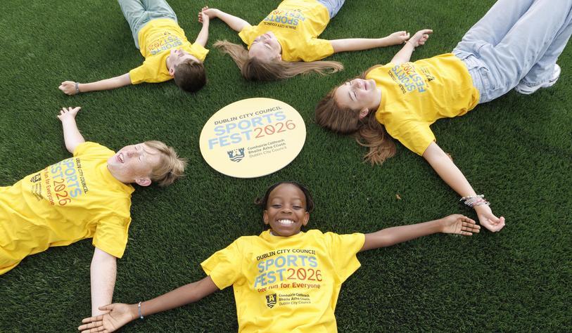 Five children lie on a sports field in a circle around a Dublin City Council SportsFest 2026 sign, smiling and wearing yellow SportsFest T-shirts.