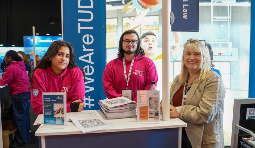 Three TU Dublin representatives stand at an information booth, with course brochures and promotional materials displayed on the table.