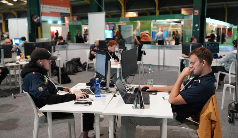Two people sit at a shared table using laptops and computer monitors in a busy indoor event or exhibition space, with other attendees and booths visible in the background.