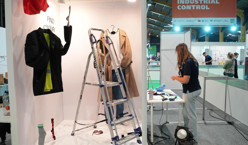 A trade show booth displays workwear, including a suit jacket and protective clothing, beside a step ladder and tools. A worker stands at a nearby workstation, with other exhibitors and attendees visible in the exhibition hall.