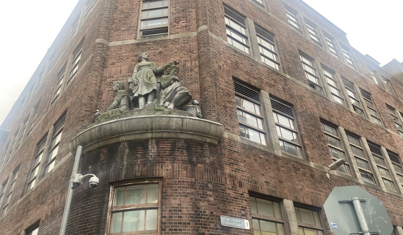 Low-angle view of a historic red-brick building featuring a stone sculptural group mounted above the entrance, with rows of large windows.