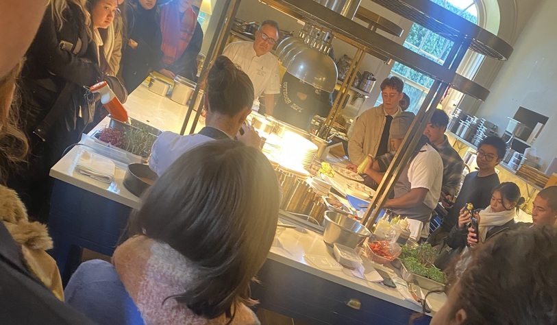 Students observe a chef demonstrating food preparation at a serving counter, with ingredients and kitchen equipment displayed as part of a culinary tour.