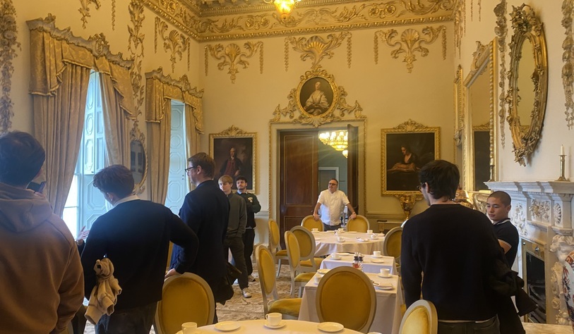 Students visit an ornate historic dining room featuring gilded decorations, portraits, chandeliers and formally set tables.