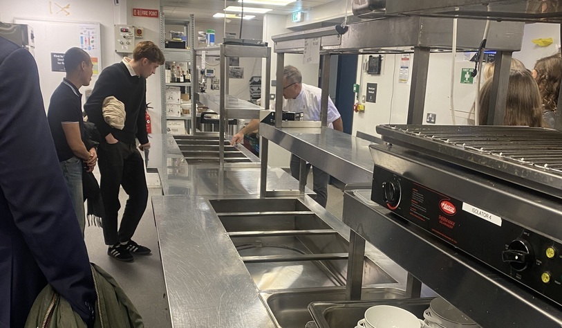 Students tour a commercial kitchen, observing a staff member working behind stainless-steel food preparation and serving counters.