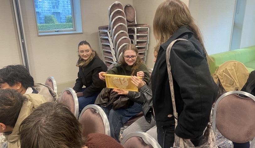 Students examine a framed sample of honeycomb during a visit, while seated in a meeting room with stacked chairs in the background.