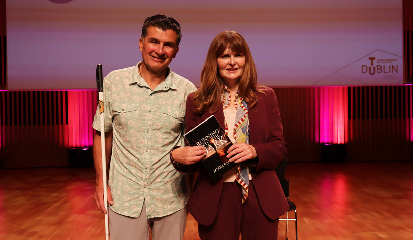 Two people pose together on a stage, with one holding a white cane and the other holding a book titled “Running the ….” Pink and red stage lighting illuminates the polished wooden floor behind them.