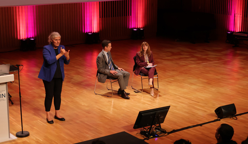 A woman speaks into a microphone on a concert hall stage while two seated panelists listen. The stage has a polished wooden floor and is illuminated by pink and red lighting.