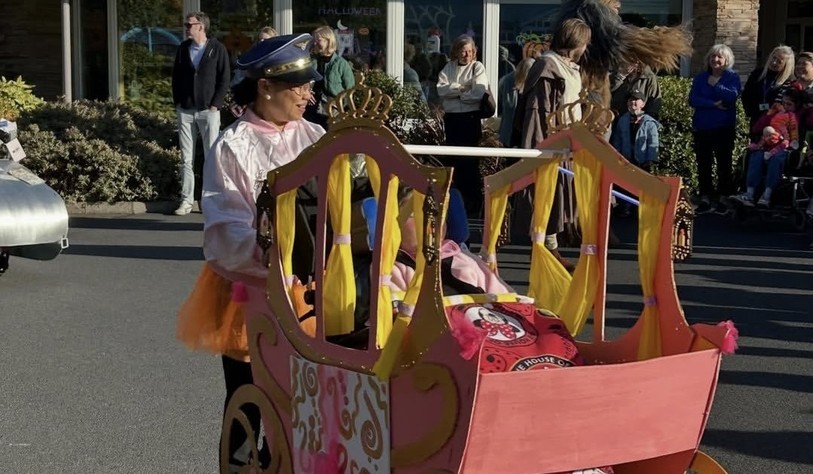 A participant dressed in a pink outfit and hat pushes a colourful homemade carriage decorated with gold crowns and pink and yellow details.