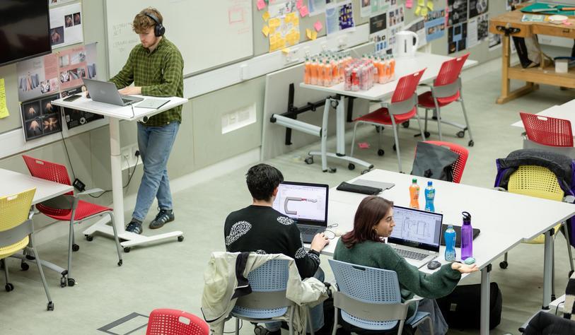 A modern design studio with students working on laptops at large tables, while another student wearing headphones works at a standing desk. Colorful chairs, project materials, and posters fill the room.