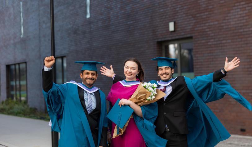 Three graduates celebrate outside a university building, smiling and waving while holding a bouquet of flowers and a graduation cap.