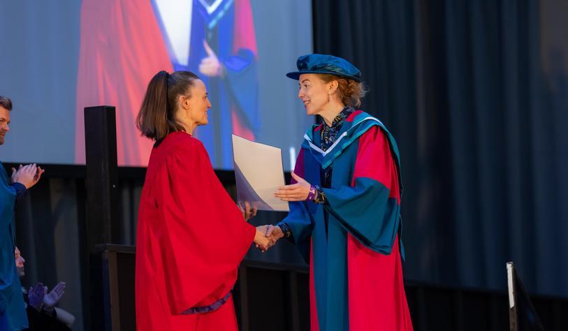 A graduate in a blue and turquoise academic gown receives a certificate and shakes hands with a university representative wearing red academic robes during the graduation ceremony.