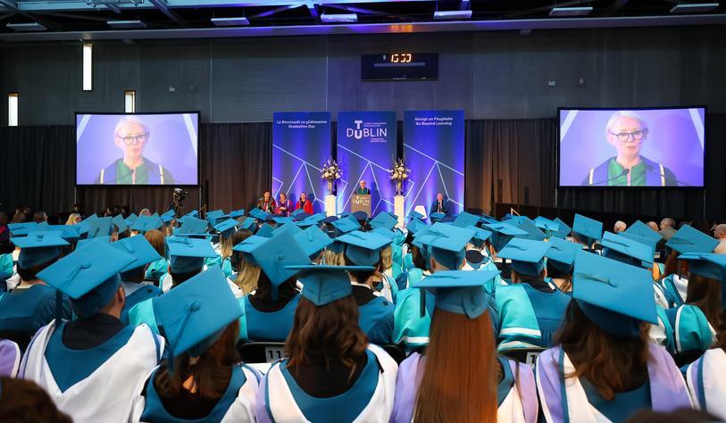 Graduates wearing turquoise mortarboards and academic gowns attend a Technological University Dublin graduation ceremony, watching the proceedings on stage and on large screens.