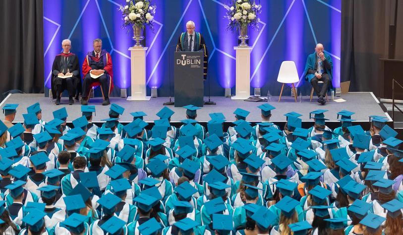 University officials in academic robes sit on stage as a speaker addresses graduates seated in rows of turquoise mortarboards during a TU Dublin graduation ceremony.