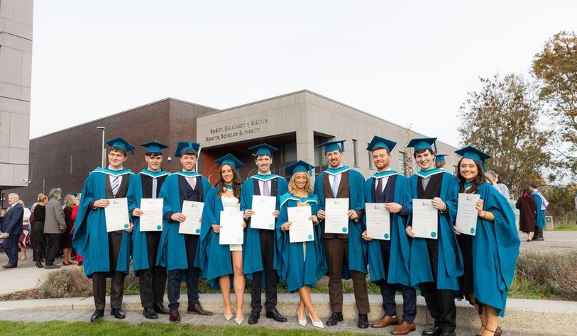 A group of graduates in blue and turquoise academic gowns and mortarboards pose outside TU Dublin’s Sports, Science and Health building, holding their graduation certificates.