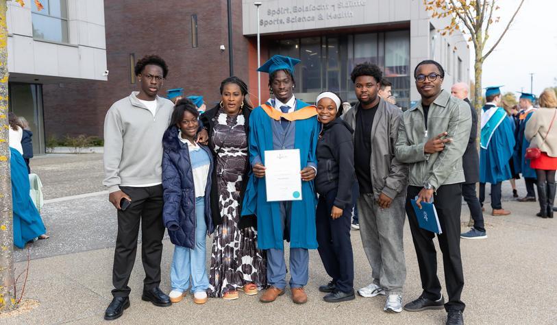 A graduate in a blue academic gown and mortarboard celebrates with family and friends outside TU Dublin’s Sports, Science and Health building, proudly holding a graduation certificate.