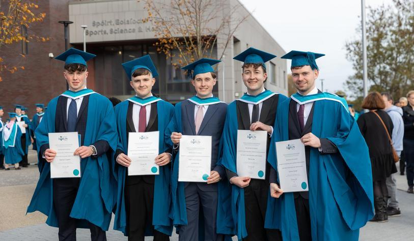 Five graduates in blue and turquoise academic gowns pose together outside TU Dublin, proudly displaying their graduation certificates.