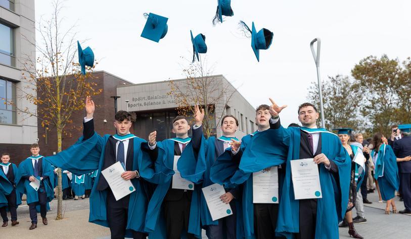 Four graduates in blue and turquoise academic gowns celebrate by throwing their mortarboards into the air outside a TU Dublin campus building.