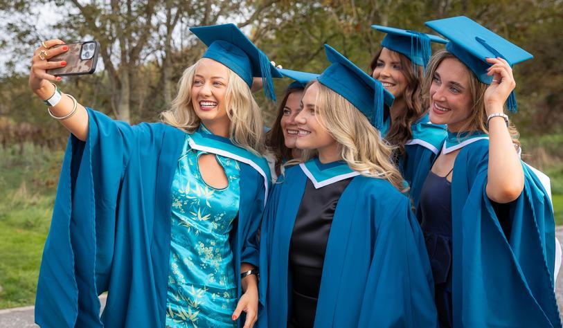 Four graduates in blue academic gowns and mortarboards smile together while taking a selfie outdoors during graduation celebrations.
