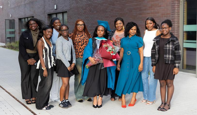 A graduate in a blue academic gown poses with family and friends outside a TU Dublin building, celebrating her graduation with a bouquet of flowers.