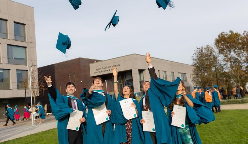 A group of graduates in blue academic gowns celebrate outdoors, throwing their mortarboards into the air while holding their graduation certificates.