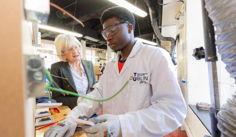 An apprentice demonstrates equipment in a workshop while wear a lab coat and protective eyewear