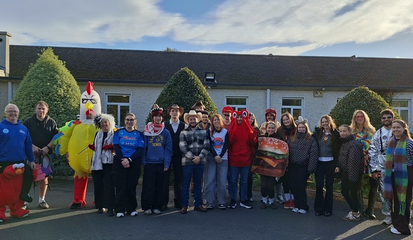 A group of staff and participants pose together outdoors in Halloween costumes, including a giant chicken, Elmo, a hamburger and a variety of other colourful characters.