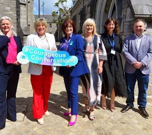 Seven adults standing outdoors in a cobblestone courtyard between stone buildings, posing together and holding a large turquoise sign that reads “#Courageous Conversations.” The group is dressed in business or smart-casual clothing, and the scene is lit by bright sunlight with architectural details and a tree visible in the background.