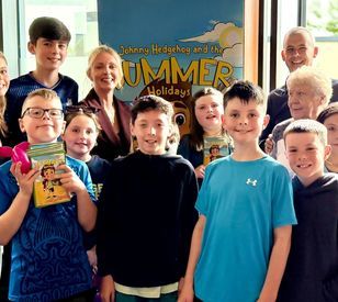 group of children and adults pose together indoors, with several children holding copies of Johnny Hedgehog and the Summer Holidays. One child holds a balloon animal, and a colourful poster for the book is visible in the background.