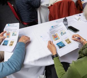 Several people sit around a table reviewing printed materials, with smartphones and papers on the table. Two people in the foreground hold illustrated information sheets.