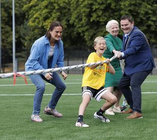 A young boy in a yellow T-shirt takes part in a tug-of-war on a sports field, supported by three adults, including two people holding the rope alongside him.
