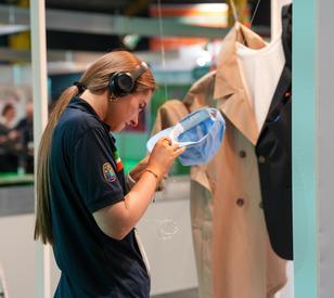 A student wearing a headset examines a blue garment or fabric item in a clothing store.