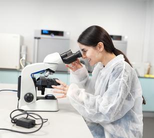 A student wearing a protective lab coat looks through a microscope in a modern science laboratory during a practical experiment.