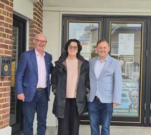 Three people standing outside a building entrance, smiling at the camera. A man in a blue jacket stands on each side of a woman wearing a dark coat.