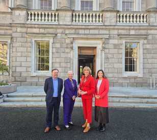 Four adults stand together outside a grand historic stone building, posing for a group photo. Three women wear colorful suits, while a man wears a dark suit with a blue shirt. The building features large sash windows, stone steps, decorative balustrades, and potted plants.