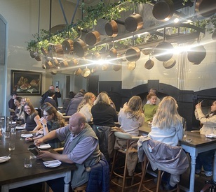 Students gather around communal tables in a café, beneath hanging planters and pendant lights.