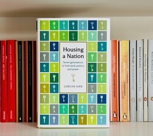 A bookshelf featuring Housing a Nation: Seven Generations of Irish Land, Politics and Power by Lorcan Sirr at the center, with a colorful key-pattern cover.
