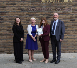 Four people stand outside a university building, with a brick wall and university signage behind them. One woman holds a certificate or document, while the others stand beside her smiling.
